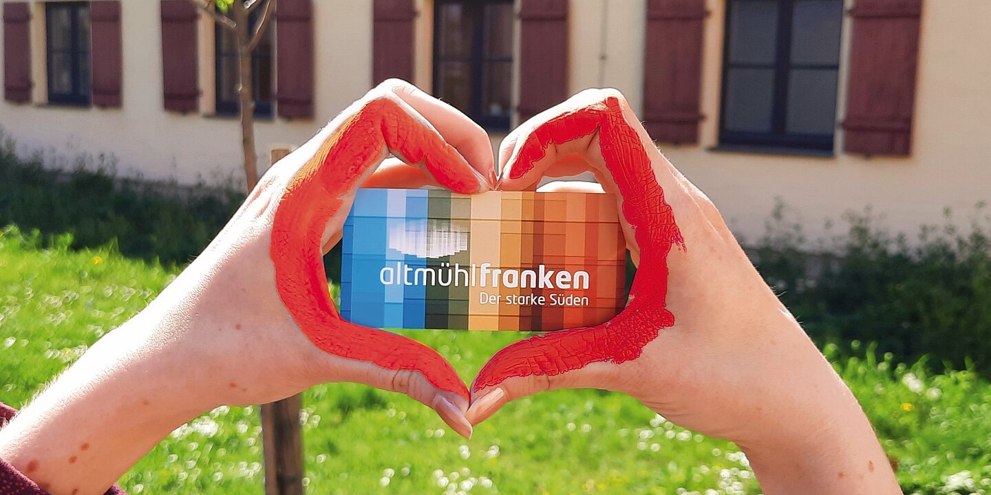 Zwei Hände die aus den Fingern ein Herz formen. Dazwischen eine Visitenkarte mit dem Altmühlfranken-Logo und dem Schriftzug Altmühlfranken. Die beiden Zeigefinger, die das Herz bilden sind mit roter Farbe bemalt. Im Hintergrund sieht man eine Wiese und einen kleinen Baum.